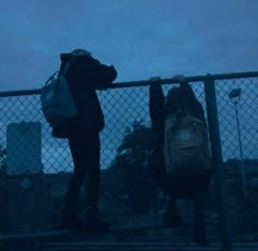 two people standing next to each other with backpacks on their backs and one holding onto the fence