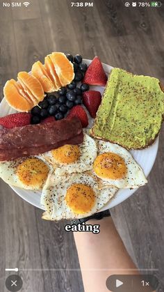 someone holding up a plate with eggs, bacon, fruit and avocado on it