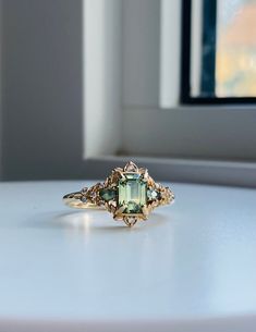 a ring with an emerald colored stone in the center on a white table next to a window