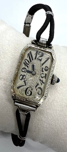 "This scarce lady's Art Deco watch features the coveted dial of exploding numbers.  By Belais, the case is 18K white gold, completely engraved, and is in perfect condition.  I don't know how much of the design on the back of the case was custom, but someone went all out in monograming.  The crown is set with a sapphire glass cabochon.  The black cord band with an Art Deco design on the clasp may or may not be original--the clasp mechanism is stamped with a patent number that dates it to 1937.  If the watch itself dates earlier, then the band is a later addition.  Judging the band, case, and crystal, I would guess that the watch had never been worn.  The dial with the fabulous exploding numbers does show some oxidation at the bottom, which isn't as bad in person as it looks in the photos. White Gold Automatic Watches For Anniversary, Timeless Engraved Watches For Anniversary, Collectible Timeless White Gold Watches, Art Deco Style Polished Jewelry And Watches For Anniversary, Art Deco Silver Jewelry And Watches For Anniversary, Timeless White Gold Collectible Watches, Engraved Watch With Rectangular Dial For Anniversary, Elegant Collectible Watch With Rectangular Dial, Engraved Watches With Rectangular Dial For Anniversary