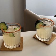 two glasses with cucumber garnish sit on a table next to each other