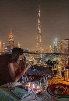 two people sitting at a table with wine glasses and plates in front of the city skyline