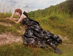 a woman laying on top of a lush green field next to tall grass covered hillside