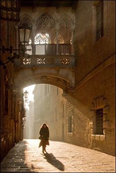 a woman is walking down an alleyway under a bridge in the city with sunlight streaming through it