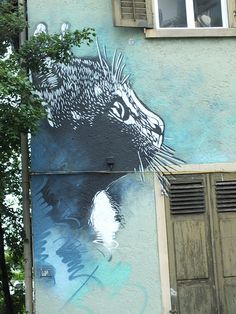 a cat painted on the side of a building with shutters open and trees in front of it