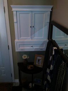 a white cabinet sitting on top of a wooden table next to a doorway with clothes hanging from it