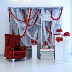 a red chair sitting in front of a window covered in sheer curtains and flowers next to a white table