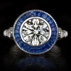 This chic Art Deco style ring features a lively and substantial 1.80ct diamond surrounded by a glamorous ring of royal blue natural sapphires. Certified by GIA with Excellent grades in all finishing categories, the diamond has fantastic brilliance! In a classic target ring design, the diamond is encircled by a rich blue border of sapphires. The effect is an air of chic Art Deco sophistication! It is an excellent choice for an elegant and historied engagement or right hand ring!The 1.80ct diamond Art Deco Gia Certified Sapphire Ring As Gift, Art Deco Sapphire Ring With Vvs Clarity, Art Deco Gia Certified Sapphire And Diamond Ring, Gia Certified Blue Art Deco Jewelry, Excellent Grades, Target Ring, Gia Certified Square-cut Sapphire Jewelry, K Color Diamond, Engagement Ring Sapphire