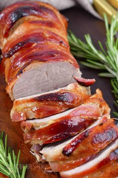 ham wrapped in bacon sitting on top of a wooden cutting board next to rosemary sprigs