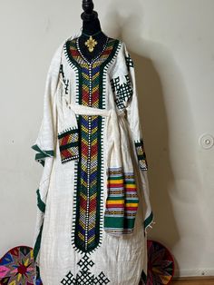 Ethiopian and Eritrean traditional dress (habesh kemis gonder kemis)garment featuring colorful and intricate embroidery. This style is often seen in Ethiopian or Eritrean cultural clothing, known as "Habesha kemis." The dress is typically worn by women for special occasions and celebrations. The garment in   likely  white cotton or linen and adorned with detailed patterns along the neckline, cuffs, and front panel, reflecting the rich textile heritage of the region. Traditional Ceremonial Sets With Woven Motifs, Traditional Sets With Weaving Work For Ceremonies, Traditional Kaftan With Multicolor Embroidery, Traditional Embroidered Kaftan With Traditional Patterns, Traditional Kaftan With Multicolor Embroidery And Patterns, Traditional Dresses With Multicolor Embroidery And Woven Motifs, Festive Habesha Kemis With Traditional Patterns, Traditional Long Sleeve Habesha Kemis For Festivals, Traditional Green Dabka Kaftan