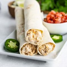 two burritos on a white plate with salsa and peppers in the bowl behind them