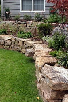 a stone wall in front of a house with grass and flowers on the ground next to it