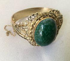 "Gold tone engraved/embossed floral design hinged cuff/clamper with filigree and saw tooth bezel. Excellent condition. Very minor rubbing wear on the inside of the bracelet. Raised applied dimensional oval design. Green mottled Peking glass oval cabochon in center of the bangle. Beautifully signed on the hinge. Nice chain guard. Very minor wear. Beautiful pierced work filigree. This bangle is a cut above other Whiting and Davis hinged bracelets. 1 3/4\" x 1/2\" band (width tapered) 44.5 grams wt Antique Hinged Cuff Bracelet Bangle, Antique Hinged Bangle Cuff Bracelet, Antique Hinged Cuff Bracelet, Vintage Hinged Bangle For Formal Occasions, Ornate Hinged Cuff Bangle Bracelet, Vintage Formal Bangle With Intricate Design, Formal Vintage Bangle With Intricate Design, Vintage Round Filigree Bangle, Vintage Filigree Bangle