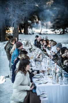 a group of people sitting at a long table with wine glasses on it in the snow