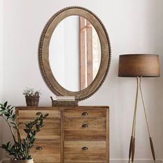 a wooden dresser with a round mirror on top of it next to a lamp and potted plant