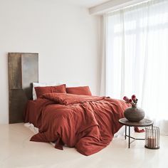 a bed with red sheets and pillows in a white room next to a table with flowers on it