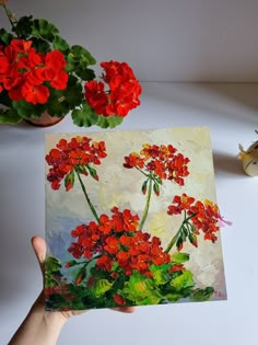 a painting of red flowers is being held by someone's hand next to a potted plant