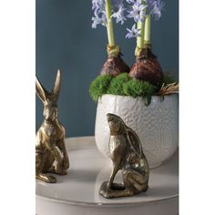 two gold rabbit figurines sitting next to each other on a white plate with purple flowers in the background