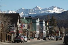 cars are parked on the street in front of buildings with snow capped mountains behind them