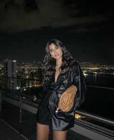 a woman standing on top of a roof next to a city at night with her hands in her pockets