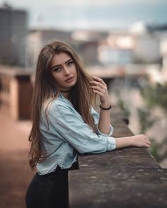 a woman with long hair leaning on a wall looking at the camera while wearing black pants and a blue shirt