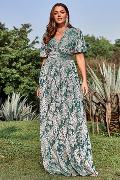 a woman wearing a green and white floral print dress standing on grass in front of trees