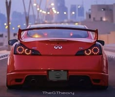 the rear end of a red sports car driving down a street at night with city lights in the background