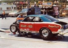 an old race car being worked on in a parking lot with other cars and people