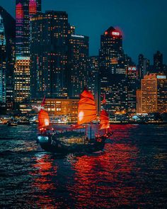 a sailboat with red sails in front of the city skyline at night, as seen from across the water