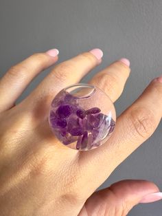 This beautiful ring combines elegance and originality with its unique 27mm glass capsule design. This ring captures the beauty of nature with its liquid filling and amethyst natural stones carefully embedded into the inside of the capsule, adding a subtle yet impactful sparkle. Amethysts is known for its spiritual and calming properties which makes this the best fashion accessory and a symbol of serenity and elegance. The ring band material is designed in transparent resin which allows the capsule and amethyst stones to shine in all their splendor while remaining comfortable and light in your hand. This ring is perfect for special occasions or to add a touch of sophistication to your everyday style. Turn every day into a magical experience by wearing this glass capsule ring with amethyst s Modern Resin Rings For Gifts, Unique Clear Crystal Ring For Gift, Unique Clear Crystal Ring As A Gift, Clear Crystal Ring Gift, Clear Crystal Ring For Gift, Unique Clear Resin Ring, Unique Resin Crystal Ring Gift, Unique Clear Rings, Unique Clear Round Rings