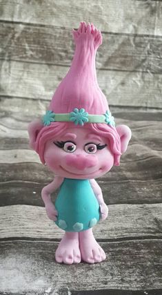 a pink troll figurine wearing a blue dress and green headband on top of a wooden table