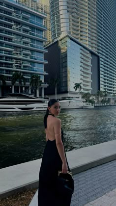 a woman in a black dress is walking by the water with buildings and boats behind her