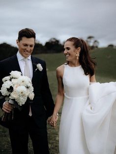 a bride and groom holding hands walking through the grass with white flowers in their hand