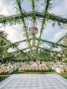 an outdoor wedding venue with greenery and chandeliers on the ceiling, surrounded by white tables and chairs