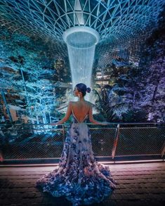 a woman standing in front of a waterfall with her back to the camera, looking at it