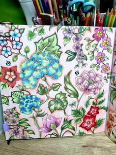 an open notebook with colorful flowers on it and pencils next to it in front of some pens