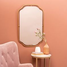 a pink chair sitting next to a table with a vase and mirror on top of it