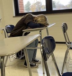 a person sitting at a desk with their head on the back of another chair, in front of a window