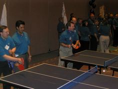 two men are playing ping pong in a large room with other people standing around