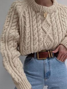 a woman wearing a sweater and jeans is holding her hands in her pockets as she wears a belt