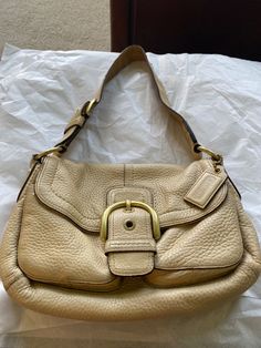 This Coach shoulder bag is a timeless piece that will elevate any outfit. The butter cream leather exterior is accented with a beautiful buckle and magnetic closure. The bag measures 11 inches in width, 8 inches in height, and 5 inches in depth, making it the perfect medium-sized accessory. The interior is lined with beige fabric and comes with a bag charm accessory. The handle and strap are made of brass and leather, adding to the durability and style of the bag. This vintage piece is from the Coach product line and has a solid pattern that is perfect for any occasion. Beige Bag Outfit, Leather Bag Outfit, Cream Bags, Beige Bag, Bag Outfit, Coach Shoulder Bag, Beige Fabric, Solid Pattern, Magnetic Closure