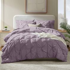 a bed with purple comforter and pillows in a room next to a plant on the floor