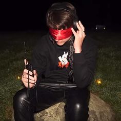 a young man sitting on top of a rock wearing a blindfold and holding two cell phones