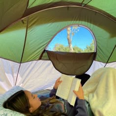 a woman laying in bed reading a book under an open green tent with trees and blue sky reflected in the window