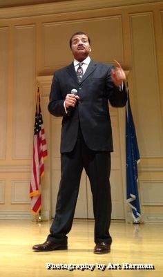 a man in a suit and tie standing on stage with an american flag behind him