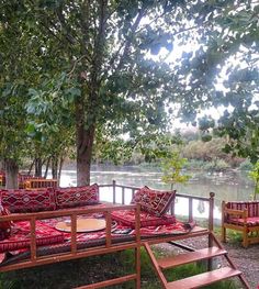 an outdoor seating area with wooden benches and red cushions on the grass near a body of water