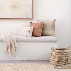 a white bench with pillows on it and a basket next to it in front of a painting