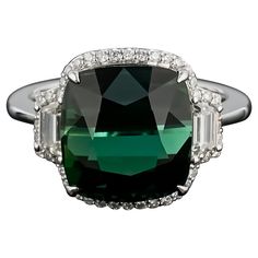 An elegant and simple cocktail ring using a 9.26 carat cushion cut Tourmaline centre stone, with 0.37 carat trapeze Diamond side stones and 0.26 carat brilliant cut diamonds sorrounding it, all set in 18K white and gold. The center stone has an ideal green color, with no inclusions and is completely transparent. The cut of the stone makes it look like a cats-eye, hence making it a very unique stone. Currently a ring size US 7, but we can resize the ring for you without additional cost Certificat Formal Fine Jewelry Cushion Cut Diamond Ring, Formal Halo Ring With Cushion Cut Center Stone, Cushion Cut Halo Ring For Formal Occasions, Formal Cushion Cut Fine Diamond Ring, Formal Cushion Cut Halo Ring With Center Stone, Dazzling Cushion Cut Halo Ring For Formal Occasions, Luxury Cushion Cut Emerald Ring With Diamond, Luxury Cushion Cut Diamond Ring For Formal Events, Luxury Cushion Cut Diamond Ring For Formal Occasions