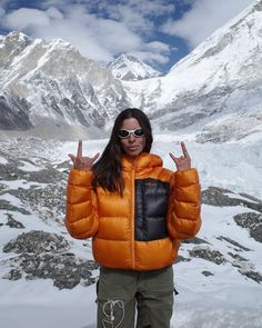 a woman in an orange jacket standing on top of a snow covered mountain giving the peace sign