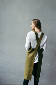 a woman standing in front of a gray wall wearing a green apron and white shirt